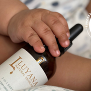 Baby's hand holding a bottle of Luyana Natural Skincare's 3-in-1 Baby Oil with a European Natural Beauty Awards Excellence badge in the corner.