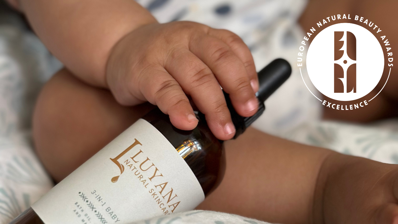 Baby's hand holding a bottle of Luyana Natural Skincare's 3-in-1 Baby Oil with a European Natural Beauty Awards Excellence badge in the corner.