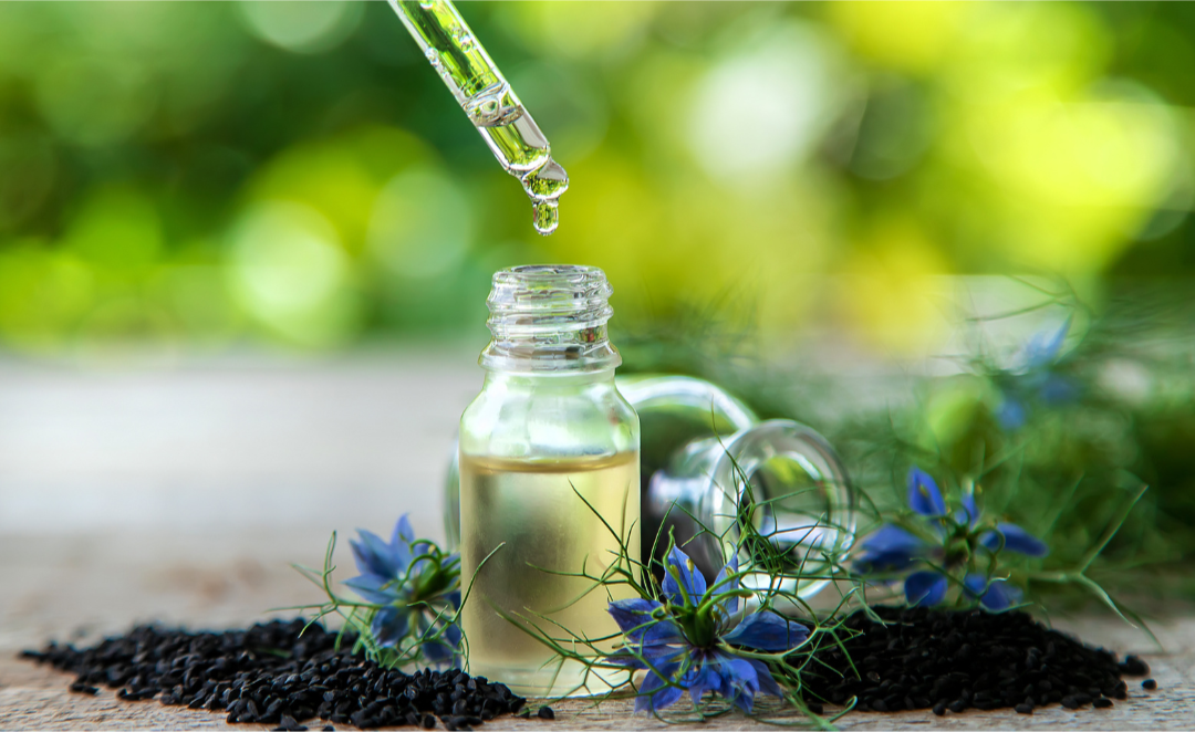 dropper hovering above bottle containing black seed oil, with nigella sativa seeds and flowers sitting on the surface around the bottle