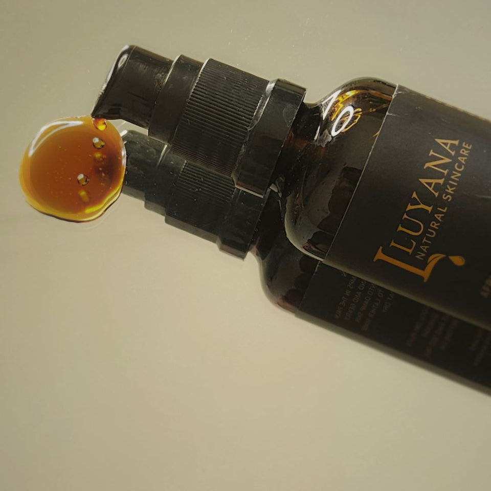 A close-up of a dark amber pump bottle labeled 'LUYANA NATURAL SKINCARE' with a glossy amber droplet resting on the pump head, set against a plain light background.