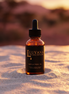 Luyana Skincare Marula Oil in a dropper bottle on sand with sunset background.